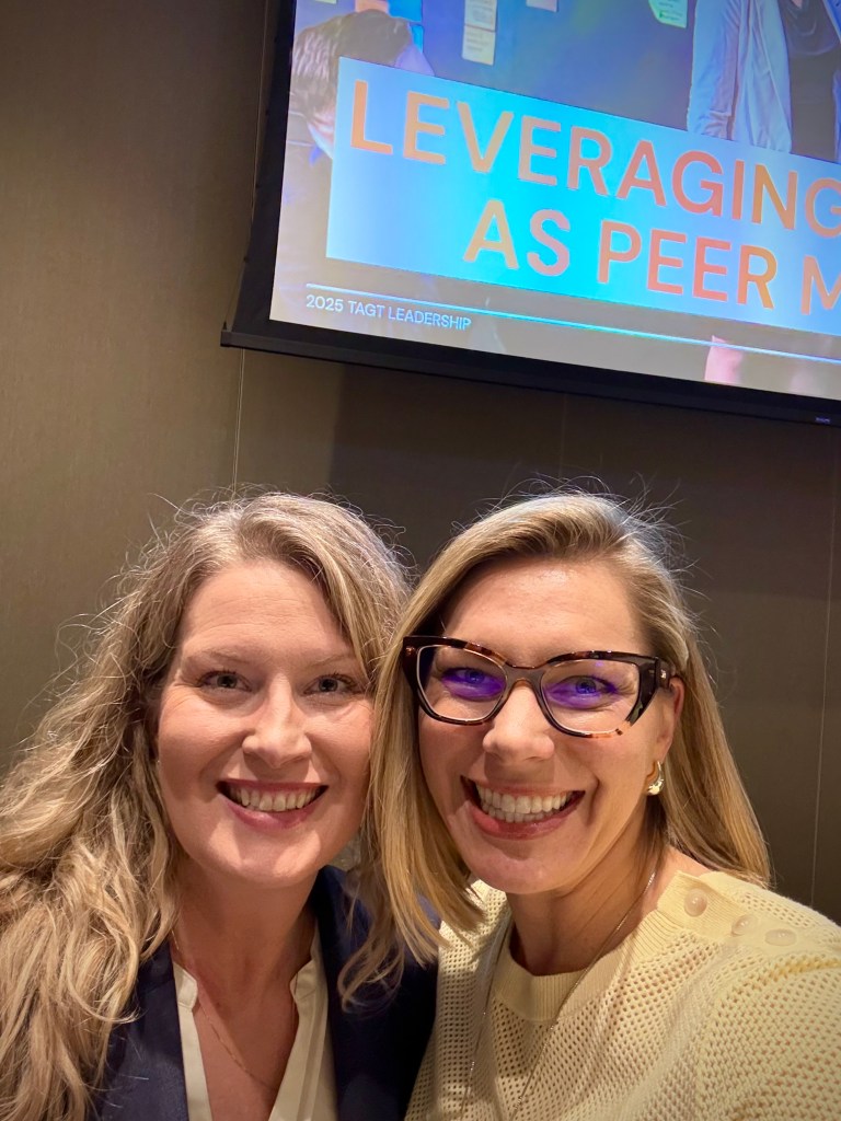 Two women smile in front of a screen projection of a slideshow